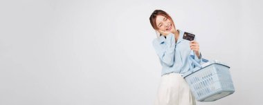 Elinde kredi kartı ve sepetle market alışverişi yapan heyecanlı Asyalı kadın, neşeli bir süpermarket ortamında finans esnekliğinin keyfini çıkarıyor. izole stüdyo beyaz arkaplan