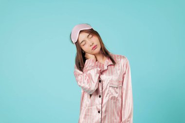 Asyalı kadın boyun ve omuz ağrısı çekiyor, uykuya ihtiyacı olduğunu hissediyor, pijama giyiyor ve maske takıyor, muhtemelen uzun uykudan sonra esniyor, vesikalık fotoğrafları stüdyo mavisi.