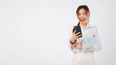 Askıda kolu kırık gülümseyen Asyalı kadın cep telefonu kullanıyor, kazada yaralanma, sosyal medyaya erişim, internet üzerinden klinik arama, telefon erişimi, izole edilmiş stüdyo beyaz arka plan