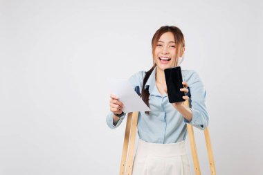 Koltuk değnekleri Asyalı hastaları destekliyor. Telefon ekranı boş ve fatura kağıdı tutuyor. Yaralanmaya rağmen, gülümsemeye ve koltuk değneklerine yaslanmaya rağmen kaza ödemesi cep telefonunda. sigorta izole beyaz arkaplan