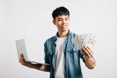Asyalı gülümseyen genç adam dizüstü bilgisayar ve para fanatiği stüdyo görüntüsü izole beyaz arka plan, mutlu yaşam tarzı işadamı dolar banknotları içinde para tutuyor.