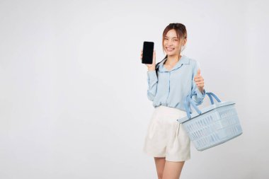 Market sepeti ve cep telefonu boş ekranı gülümseyen Asyalı kadın tarafından gösteriliyor, alışveriş anı mutlu bir şekilde süpermarkette çevrimiçi alışveriş sevincini gösteriyor. izole stüdyo beyaz arkaplan