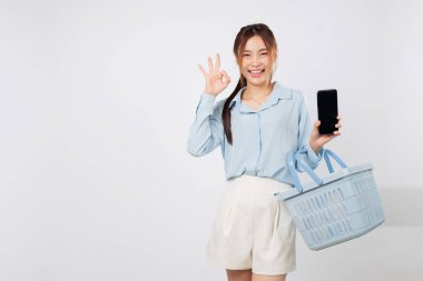 Gülümseyen Asyalı kadın sepeti ve cep telefonunu gösteriyor, alışveriş anı markette ve süpermarket sahnesinde yakalanmış. Müşteri memnuniyeti izole edilmiş beyaz arka plan