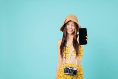 Yaz kıyafetleri içinde gülümseyen Asyalı genç bir kadın cep telefonunun boş ekranını gezintiye hazır olduğunu, eğlenceli turist yolcularının seyahat planladığını, yeni yerlere seyahat ettiğini, stüdyo mavi arka planda çekilmiş vesikalıklarını gösteriyor.