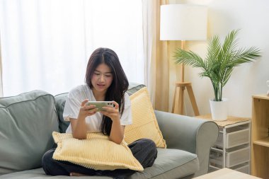 Cep telefonu kullanan Asyalı bir kadın, rahat koltukta dinlenen genç bir insan, akıllı telefondan keyif alan, oyunlar oynayan, parlak oturma odasında internette gezinen.