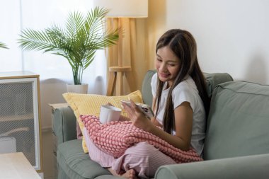 Mutlu genç bir kadın cep telefonu kullanıyor, evinde dinleniyor, akıllı telefonuyla online içeriğin tadını çıkarıyor, rahat yeşil koltukta sıcak bir içecek tutuyor, rahat yastıklarla çevrili, günlük kıyafetlerle....