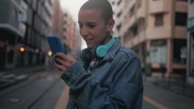 Shaved head girl using mobile smartphone while waiting for public transportation in the city street