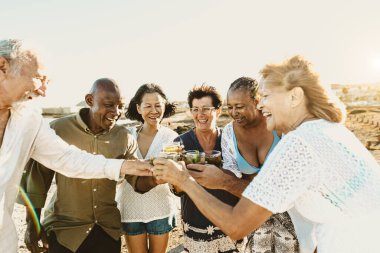 Mutlu çok ırklı kıdemli arkadaşlar gün batımında sahilde içki içerek ve mojito içerek eğleniyorlar.