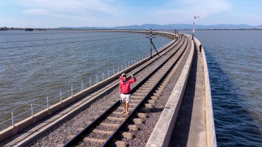 Lopburi, Thailand - January 2023 - Unidentified tourist from aerial view of an amazing travel train parked on a floating railway bridge over the water of the lake in Pa Sak Jolasid dam with blue sky 
