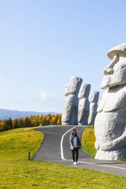 Sapporo, Hokkaido, Japonya - Kasım 2023: Gökyüzü geçmişi olan güzel bir bayan ve Buda Tepesi 'ndeki Buda heykelinin etrafında antik bir taş heykel.