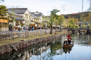 Okayama Bölgesi, Japonya - Aralık 2024: Canlı sonbahar sezonu boyunca Kurashiki Bikan Tarihi Çeyreği manzarası. Turistler geleneksel beyaz duvarlı binalar, akçaağaç ağaçları ve kanallarla dolu korunmuş Edo dönemi caddeleri boyunca yürüyorlar.