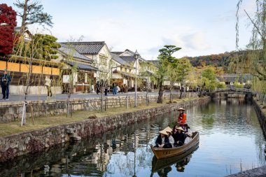Okayama Bölgesi, Japonya - Aralık 2024: Canlı sonbahar sezonu boyunca Kurashiki Bikan Tarihi Çeyreği manzarası. Turistler geleneksel beyaz duvarlı binalar, akçaağaç ağaçları ve kanallarla dolu korunmuş Edo dönemi caddeleri boyunca yürüyorlar.