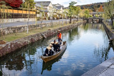 Okayama Bölgesi, Japonya - Aralık 2024: Canlı sonbahar sezonu boyunca Kurashiki Bikan Tarihi Çeyreği manzarası. Turistler geleneksel beyaz duvarlı binalar, akçaağaç ağaçları ve kanallarla dolu korunmuş Edo dönemi caddeleri boyunca yürüyorlar.