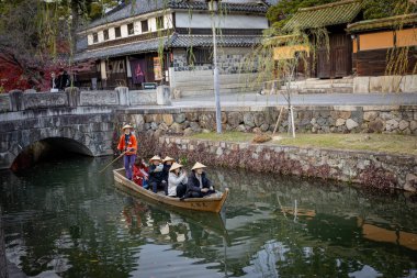 Okayama Bölgesi, Japonya - Aralık 2024: Canlı sonbahar sezonu boyunca Kurashiki Bikan Tarihi Çeyreği manzarası. Turistler geleneksel beyaz duvarlı binalar, akçaağaç ağaçları ve kanallarla dolu korunmuş Edo dönemi caddeleri boyunca yürüyorlar.