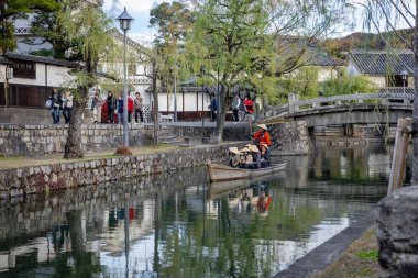Okayama Bölgesi, Japonya - Aralık 2024: Canlı sonbahar sezonu boyunca Kurashiki Bikan Tarihi Çeyreği manzarası. Turistler geleneksel beyaz duvarlı binalar, akçaağaç ağaçları ve kanallarla dolu korunmuş Edo dönemi caddeleri boyunca yürüyorlar.