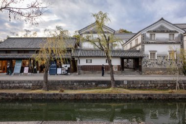 Okayama Bölgesi, Japonya - Aralık 2024: Canlı sonbahar sezonu boyunca Kurashiki Bikan Tarihi Çeyreği manzarası. Turistler geleneksel beyaz duvarlı binalar, akçaağaç ağaçları ve kanallarla dolu korunmuş Edo dönemi caddeleri boyunca yürüyorlar.