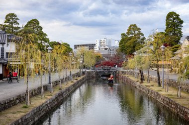 Okayama Bölgesi, Japonya - Aralık 2024: Canlı sonbahar sezonu boyunca Kurashiki Bikan Tarihi Çeyreği manzarası. Turistler geleneksel beyaz duvarlı binalar, akçaağaç ağaçları ve kanallarla dolu korunmuş Edo dönemi caddeleri boyunca yürüyorlar.