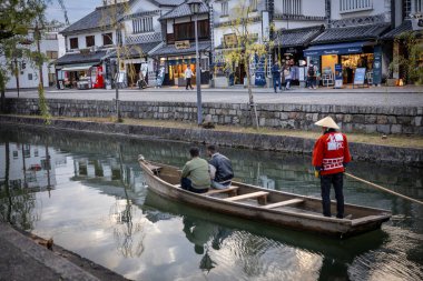 Okayama Bölgesi, Japonya - Aralık 2024: Canlı sonbahar sezonu boyunca Kurashiki Bikan Tarihi Çeyreği manzarası. Turistler geleneksel beyaz duvarlı binalar, akçaağaç ağaçları ve kanallarla dolu korunmuş Edo dönemi caddeleri boyunca yürüyorlar.