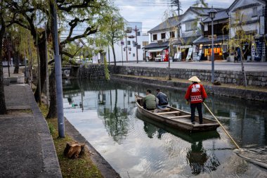 Okayama Bölgesi, Japonya - Aralık 2024: Canlı sonbahar sezonu boyunca Kurashiki Bikan Tarihi Çeyreği manzarası. Turistler geleneksel beyaz duvarlı binalar, akçaağaç ağaçları ve kanallarla dolu korunmuş Edo dönemi caddeleri boyunca yürüyorlar.