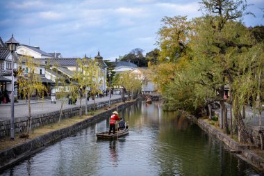 Okayama Bölgesi, Japonya - Aralık 2024: Canlı sonbahar sezonu boyunca Kurashiki Bikan Tarihi Çeyreği manzarası. Turistler geleneksel beyaz duvarlı binalar, akçaağaç ağaçları ve kanallarla dolu korunmuş Edo dönemi caddeleri boyunca yürüyorlar.