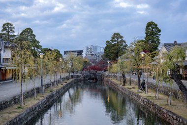 Okayama Bölgesi, Japonya - Aralık 2024: Canlı sonbahar sezonu boyunca Kurashiki Bikan Tarihi Çeyreği manzarası. Turistler geleneksel beyaz duvarlı binalar, akçaağaç ağaçları ve kanallarla dolu korunmuş Edo dönemi caddeleri boyunca yürüyorlar.