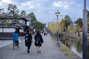 Okayama Bölgesi, Japonya - Aralık 2024: Canlı sonbahar sezonu boyunca Kurashiki Bikan Tarihi Çeyreği manzarası. Turistler geleneksel beyaz duvarlı binalar, akçaağaç ağaçları ve kanallarla dolu korunmuş Edo dönemi caddeleri boyunca yürüyorlar.