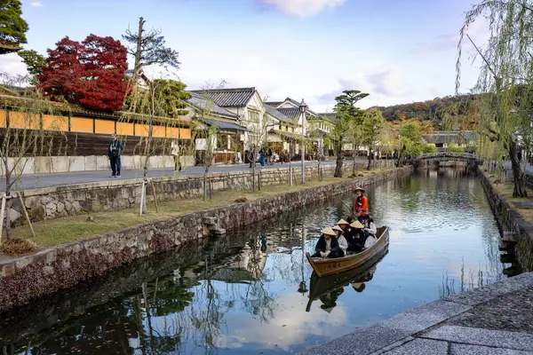 Okayama Bölgesi, Japonya - Aralık 2024: Canlı sonbahar sezonu boyunca Kurashiki Bikan Tarihi Çeyreği manzarası. Turistler geleneksel beyaz duvarlı binalar, akçaağaç ağaçları ve kanallarla dolu korunmuş Edo dönemi caddeleri boyunca yürüyorlar.