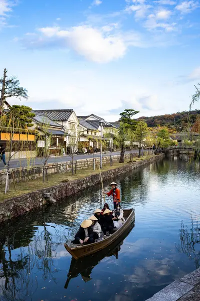Okayama Bölgesi, Japonya - Aralık 2024: Canlı sonbahar sezonu boyunca Kurashiki Bikan Tarihi Çeyreği manzarası. Turistler geleneksel beyaz duvarlı binalar, akçaağaç ağaçları ve kanallarla dolu korunmuş Edo dönemi caddeleri boyunca yürüyorlar.