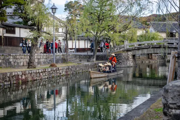 Okayama Bölgesi, Japonya - Aralık 2024: Canlı sonbahar sezonu boyunca Kurashiki Bikan Tarihi Çeyreği manzarası. Turistler geleneksel beyaz duvarlı binalar, akçaağaç ağaçları ve kanallarla dolu korunmuş Edo dönemi caddeleri boyunca yürüyorlar.
