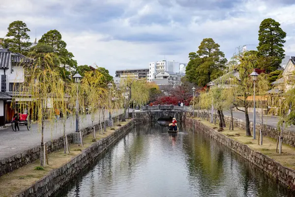 Okayama Bölgesi, Japonya - Aralık 2024: Canlı sonbahar sezonu boyunca Kurashiki Bikan Tarihi Çeyreği manzarası. Turistler geleneksel beyaz duvarlı binalar, akçaağaç ağaçları ve kanallarla dolu korunmuş Edo dönemi caddeleri boyunca yürüyorlar.