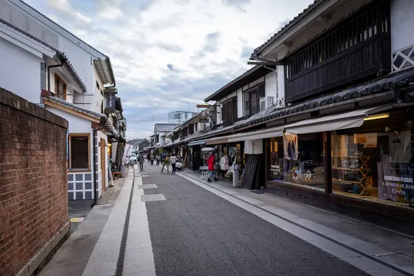 Okayama Bölgesi, Japonya - Aralık 2024: Canlı sonbahar sezonu boyunca Kurashiki Bikan Tarihi Çeyreği manzarası. Turistler geleneksel beyaz duvarlı binalar, akçaağaç ağaçları ve kanallarla dolu korunmuş Edo dönemi caddeleri boyunca yürüyorlar.