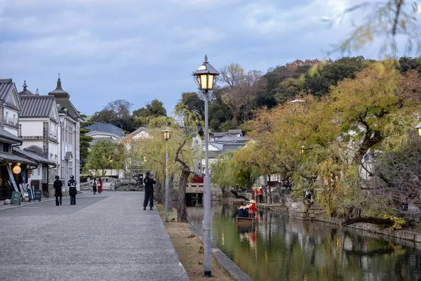 Okayama Bölgesi, Japonya - Aralık 2024: Canlı sonbahar sezonu boyunca Kurashiki Bikan Tarihi Çeyreği manzarası. Turistler geleneksel beyaz duvarlı binalar, akçaağaç ağaçları ve kanallarla dolu korunmuş Edo dönemi caddeleri boyunca yürüyorlar.