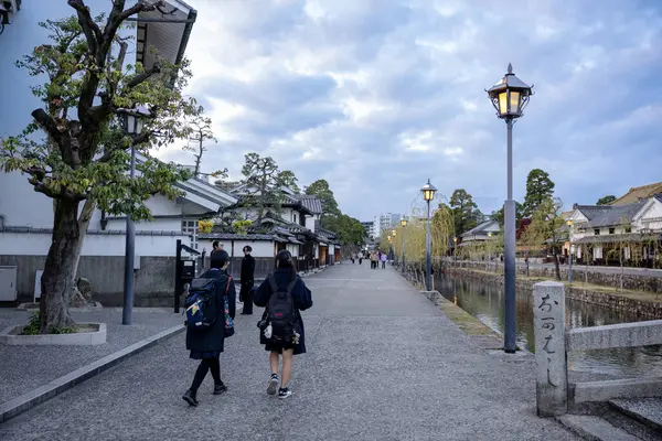 Okayama Bölgesi, Japonya - Aralık 2024: Canlı sonbahar sezonu boyunca Kurashiki Bikan Tarihi Çeyreği manzarası. Turistler geleneksel beyaz duvarlı binalar, akçaağaç ağaçları ve kanallarla dolu korunmuş Edo dönemi caddeleri boyunca yürüyorlar.