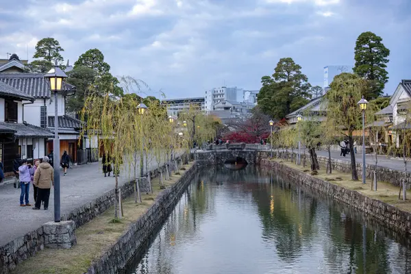 Okayama Bölgesi, Japonya - Aralık 2024: Canlı sonbahar sezonu boyunca Kurashiki Bikan Tarihi Çeyreği manzarası. Turistler geleneksel beyaz duvarlı binalar, akçaağaç ağaçları ve kanallarla dolu korunmuş Edo dönemi caddeleri boyunca yürüyorlar.