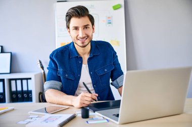 Grafik tableti ve dizüstü bilgisayarla çalışırken kameraya bakan genç tasarımcı