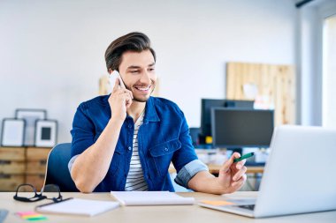 Ofiste dizüstü bilgisayarla çalışırken telefonla konuşan gülümseyen genç web geliştiricisi