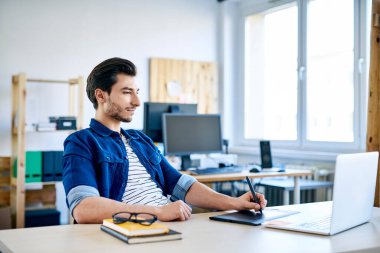Grafik tableti ve dizüstü bilgisayarla çalışan genç tasarımcı küçük ofisteki masada.