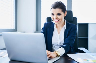 Ofisinde çalışmak için dizüstü bilgisayar kullanan gülümseyen genç bir iş kadını.