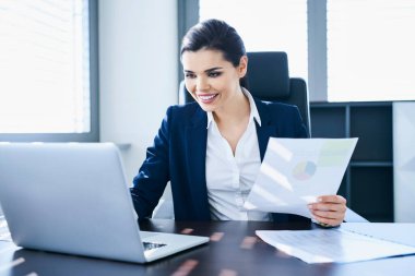 Masasında çalışan gülümseyen bir kadın notlar ve dizüstü bilgisayar kullanıyor.