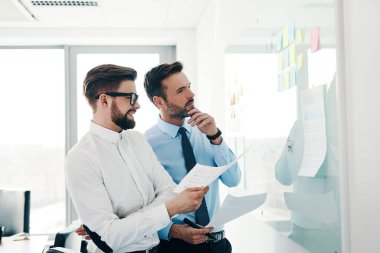 Modern ofiste birlikte çalışan iki iş adamı yeni projeyi beyaz tahtada tartışıyor.