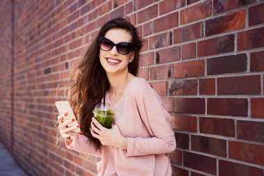 Mutlu genç kadın telefonunu kullanıyor ve ıspanaklı smoothie içiyor.
