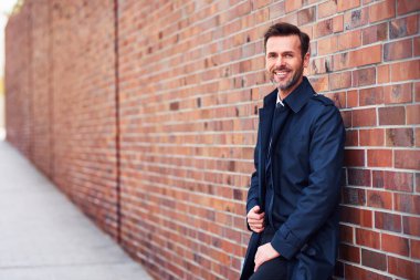 Gülümseyen orta yaşlı işadamı kameraya bakıyor ve duvara yaslanıyor.