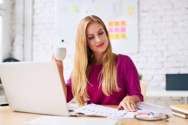 Çekici bir iş kadını modern ofisinde evrak işleriyle uğraşırken kahve içiyor.