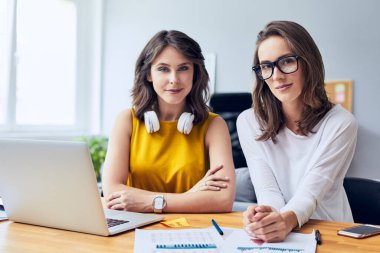 Ofiste dizüstü bilgisayarla otururken kameraya bakan iki genç girişimci.