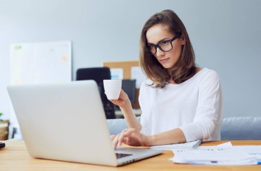Elinde kahve tutan ve modern ofiste dizüstü bilgisayar kullanan genç bir iş kadını.