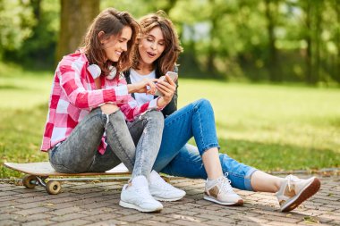Uzun panoda oturan neşeli bayan arkadaşlar akıllı telefonlara bakıyorlar.