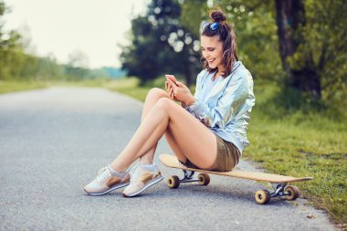 Longboard 'da oturan ve dışarıda telefon kullanan mutlu genç bir kadın.