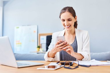 Ofiste oturan neşeli, genç bir kadın telefon kullanıyor.