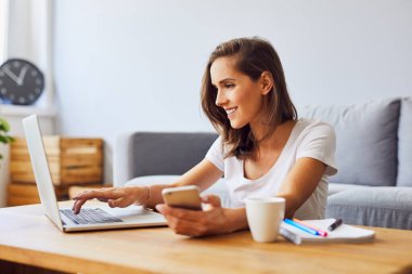 Dizüstü bilgisayar ve telefon kullanan genç bir kadın evdeki oturma odasında oturuyor.