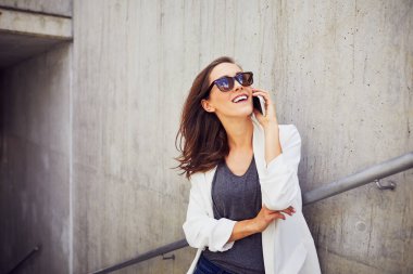 Sıradan giyinmiş genç bir iş kadını dışarıda telefonla konuşuyor.